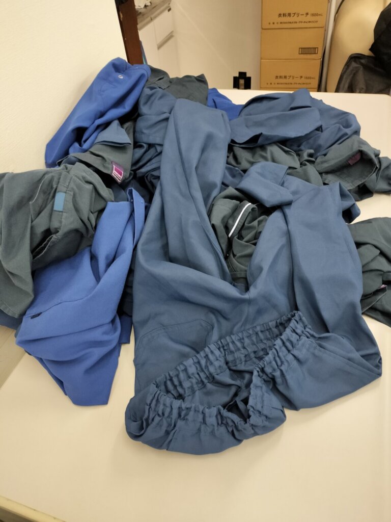 医療スクラブが大量に積まれている回収直後の状態。青・紺色の制服が混在している。